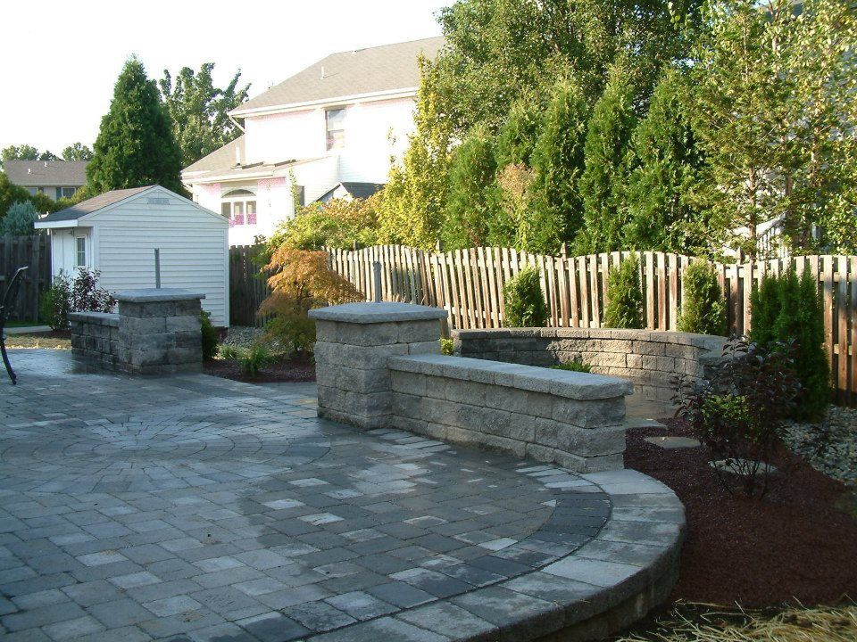 A paved backyard patio featuring stone retaining walls, pillars, and landscaping, set against a wooden fence and house. A paved backyard patio featuring stone retaining walls, pillars, and landscaping, set against a wooden fence and house.