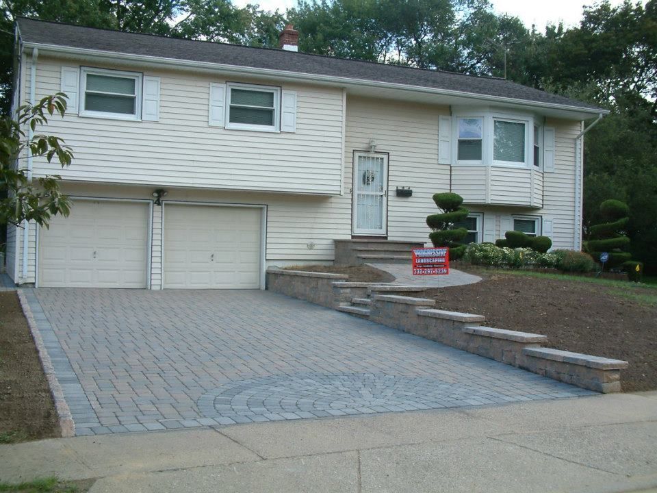 A beige split-level house featuring a paver driveway, stone retaining walls, and a new paved walkway to the front door.