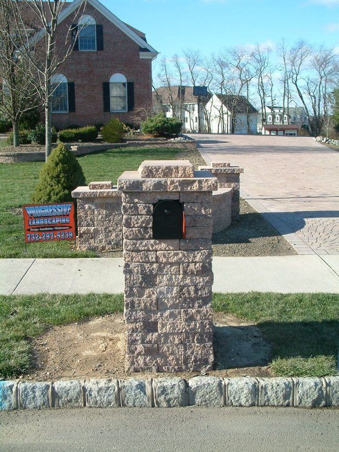 A tan, block-style stone mailbox column stands on a residential lawn in front of a brick house. A tan, block-style stone mailbox column stands on a residential lawn in front of a brick house.