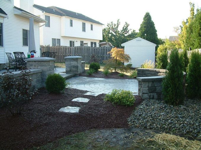 A backyard patio with stone pavers, a small retaining wall, dark mulch, stone steps, and landscaping in a suburban yard. A backyard patio with stone pavers, a small retaining wall, dark mulch, stone steps, and landscaping in a suburban yard.