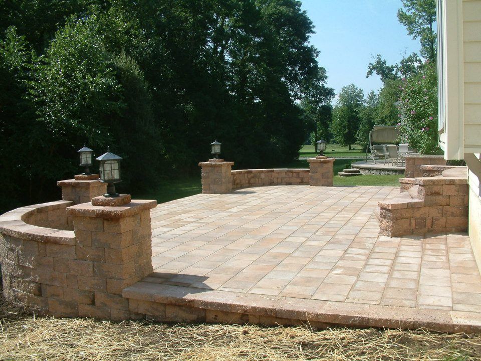 A stone patio with a low curved retaining wall, built-in pillars topped with lamps, and a stone bench, outdoors.