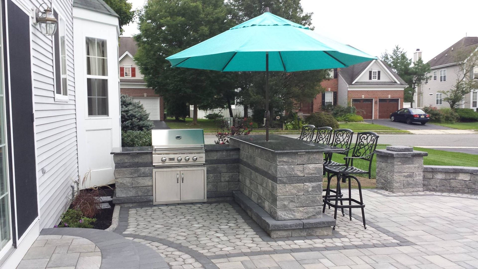 An outdoor patio kitchen with a stainless steel grill, stone bar, and a teal umbrella next to a suburban house.