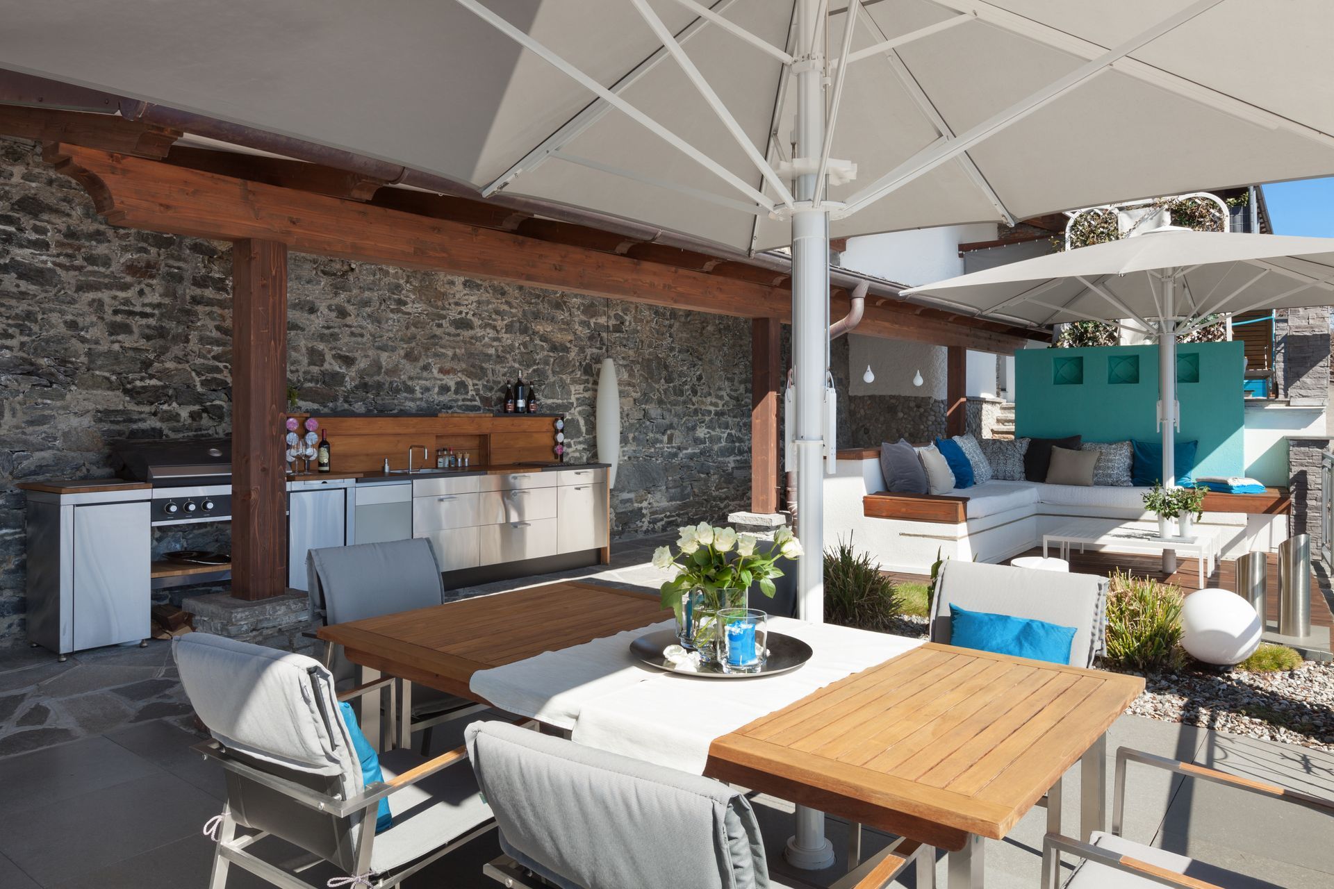 Outdoor patio dining area with wooden tables, gray chairs, and white umbrellas beside a stone wall Outdoor patio dining area with wooden tables, gray chairs, and white umbrellas beside a stone wall