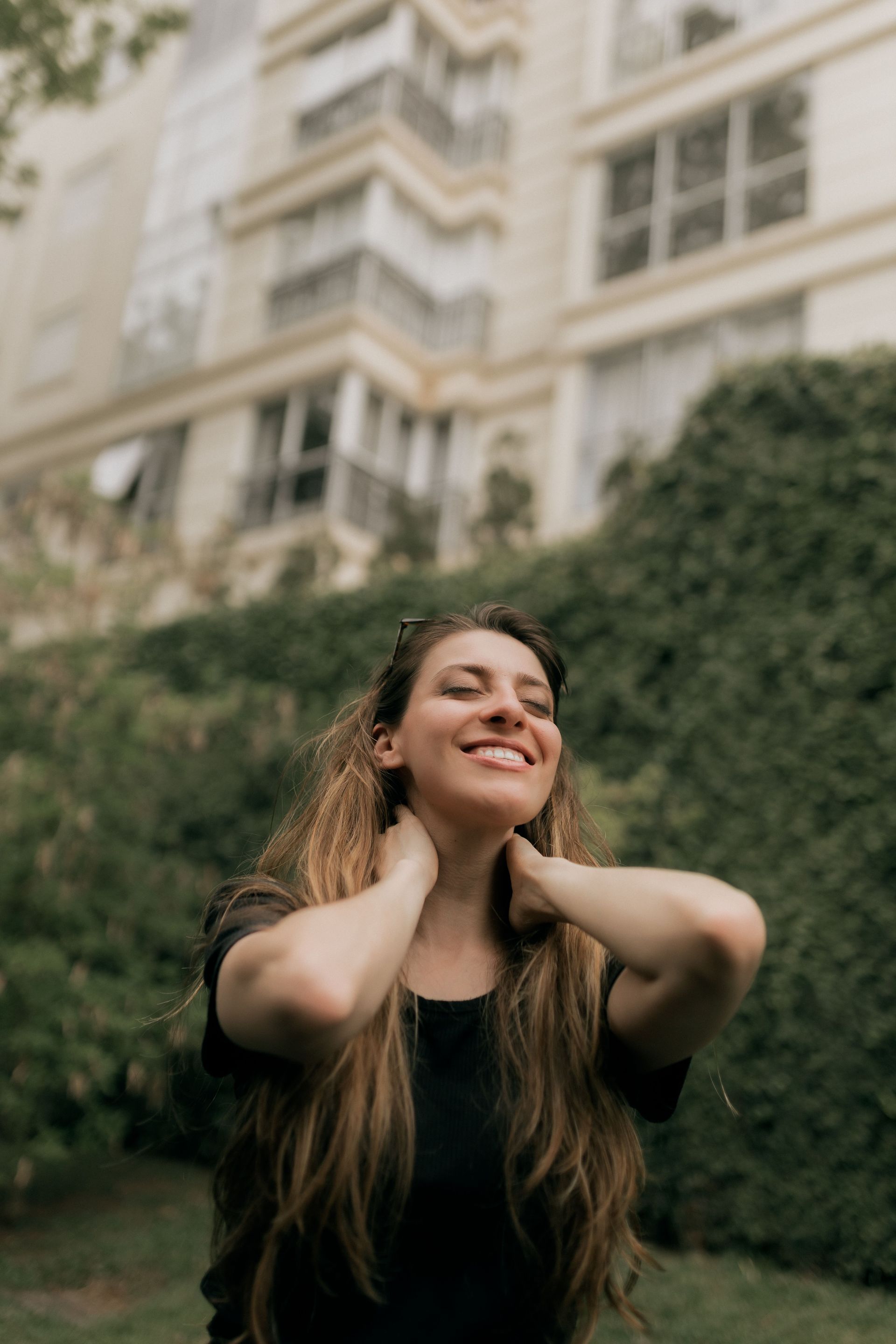 Woman smiling with hands at her neck in front of a hedge and a building