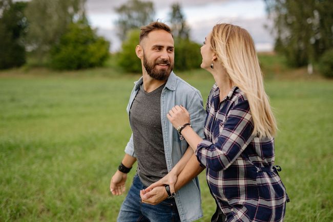 Two people walking arm in arm through a grassy park, smiling at each other