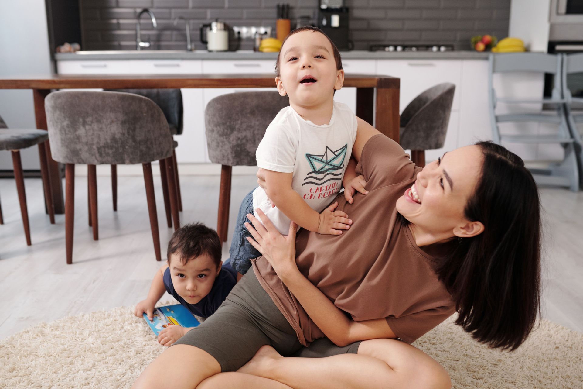 Two children play with a woman on a living room floor while a third child stands nearby.
