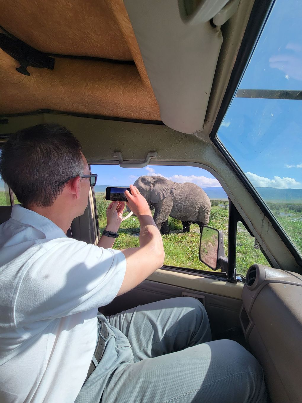 A person in a safari vehicle captures a photo of an elephant standing in a grassy field on a sunny day.
