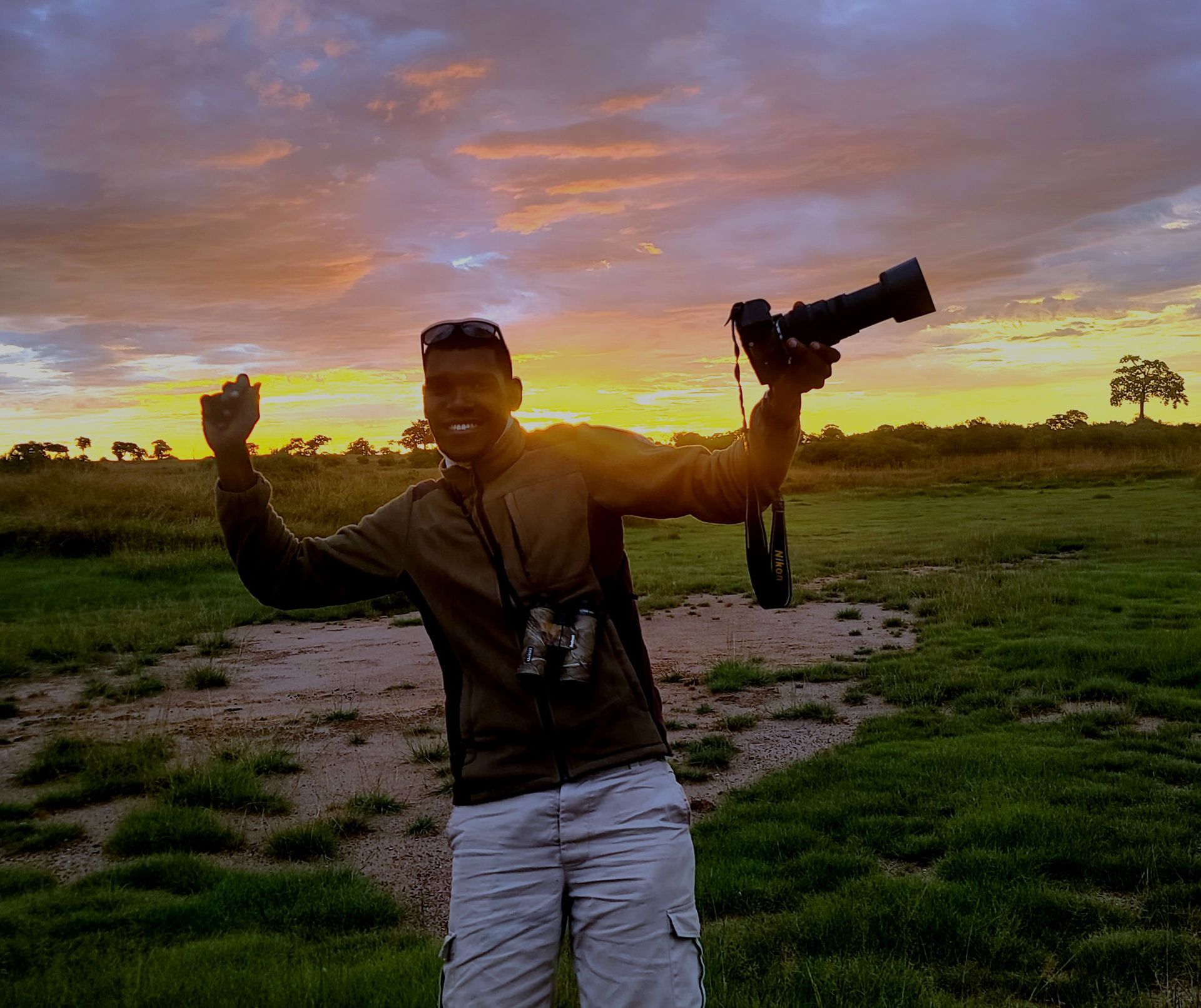A safari guide in a grassy field at sunset, smiling with arms raised, holding a camera in one hand and wearing binoculars.