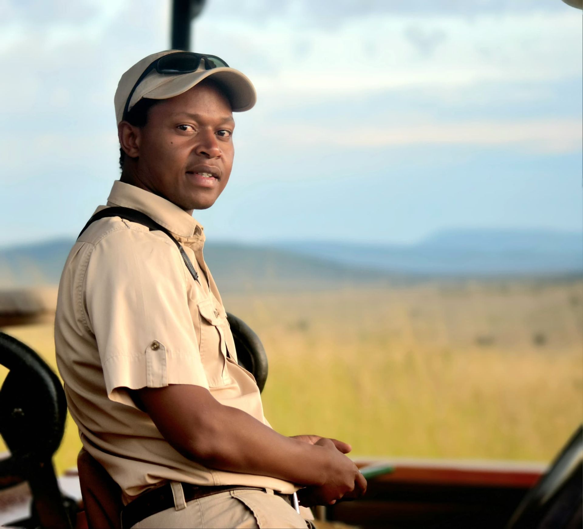 A safari guide wearing a tan uniform and cap sits in an open-air vehicle, overlooking a grassy savanna at sunset.
