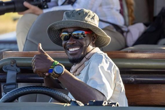 A smiling guide in a sun hat and sunglasses sits behind a steering wheel, giving a thumbs-up gesture in an open vehicle.
