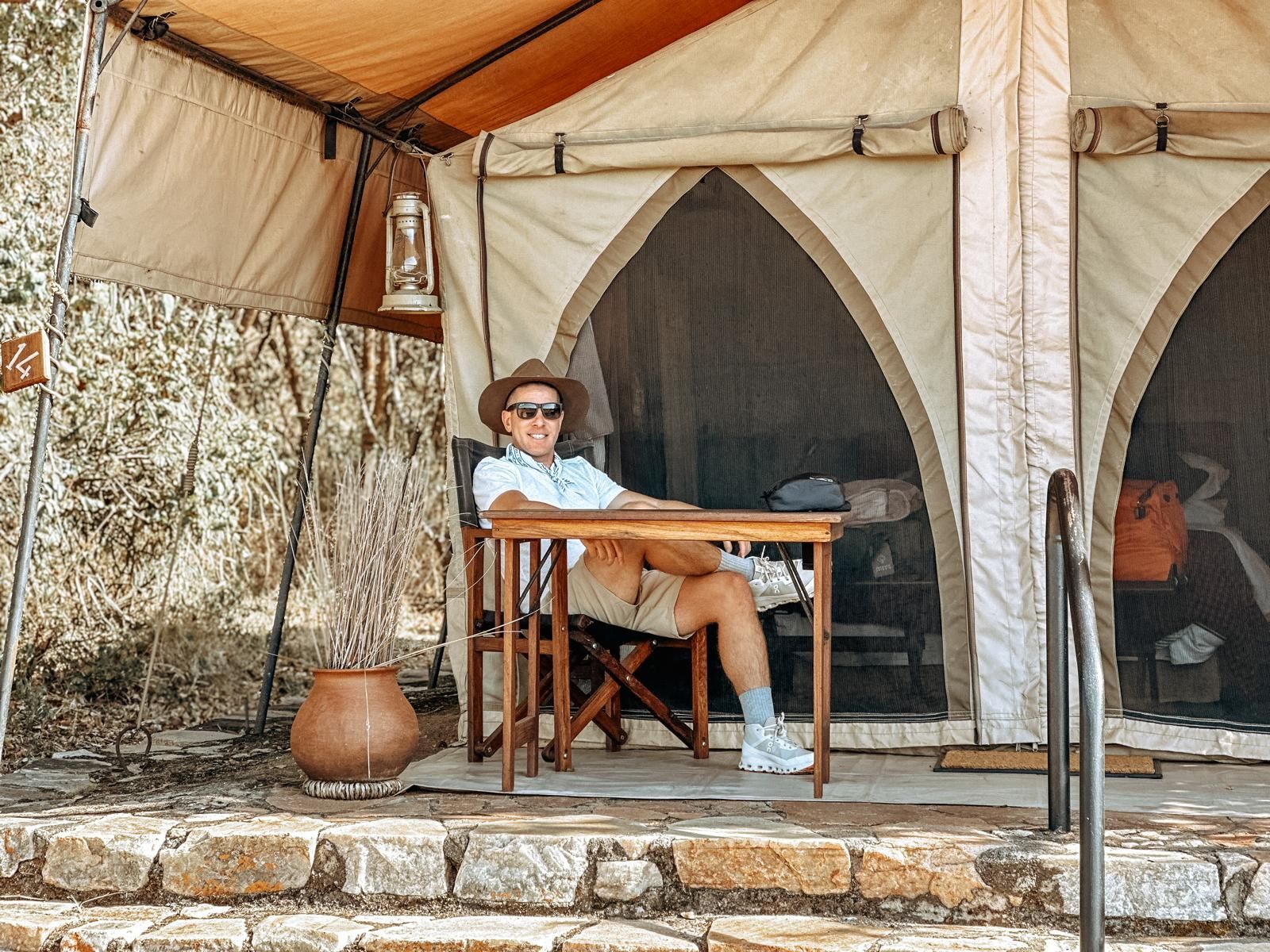 Person lounging at a table under a tan tent at a campsite, wearing sunglasses and a hat.
