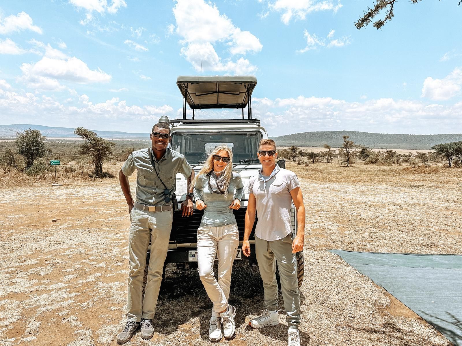 Three people stand smiling before a safari vehicle parked in a dry, open savanna landscape under a bright, sunny sky.