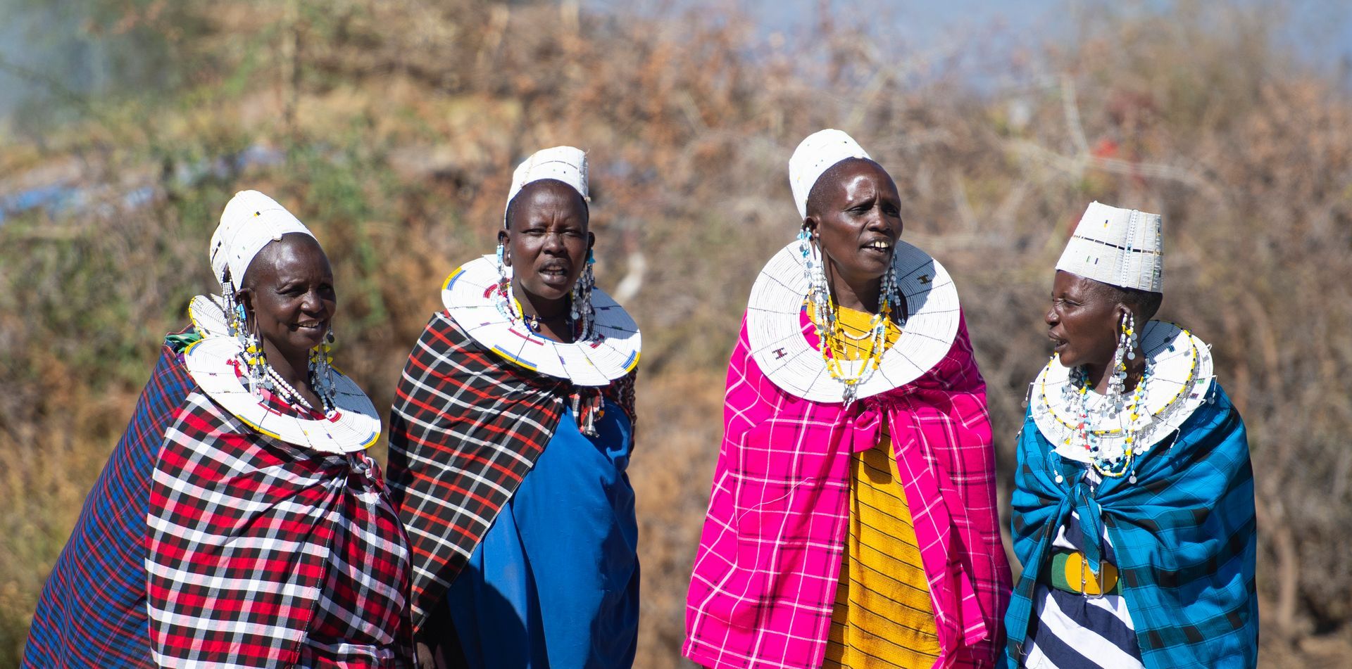 Four African women wearing white headpieces, colorful blankets, and beaded necklaces, standing together outdoors.