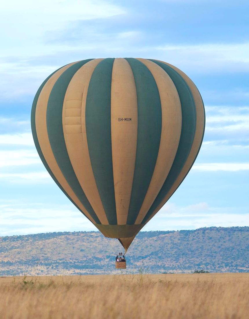 Tan and teal hot air balloon floating over dry grassland with distant hills and blue sky