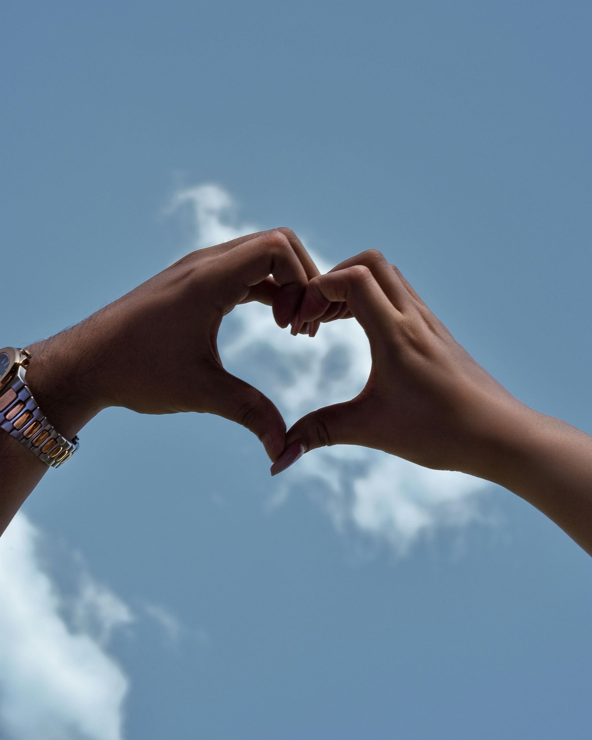 Hands forming a heart shape against a blue sky with clouds