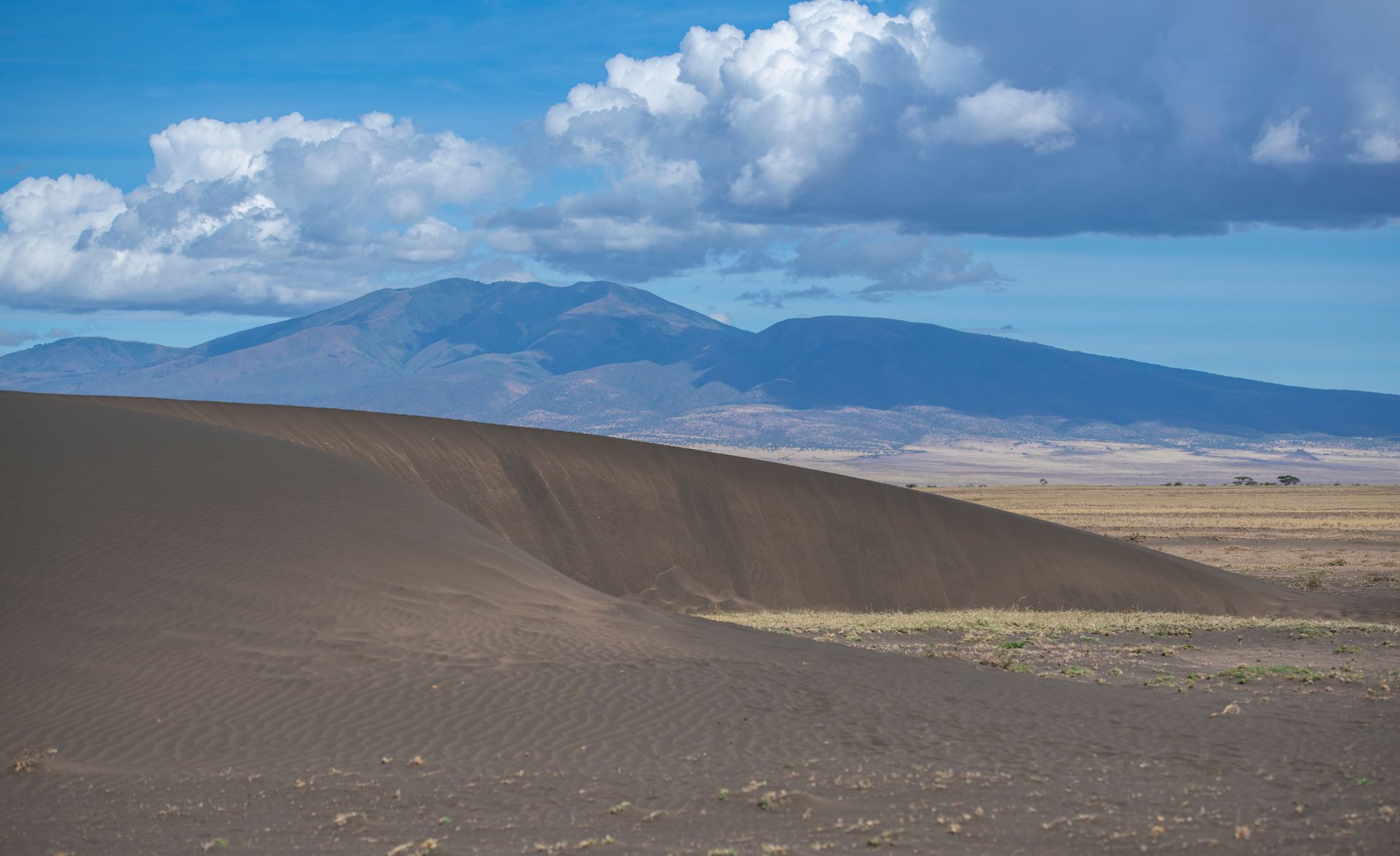 A vast, dark volcanic slope leads toward distant, hazy mountains under a blue sky with fluffy white clouds.