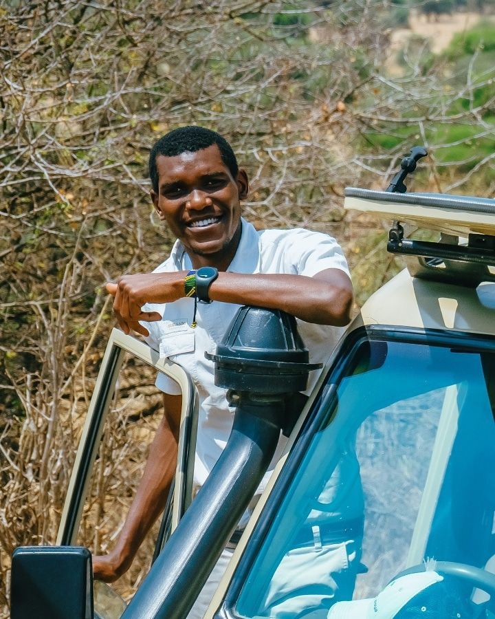 A smiling individual in a white shirt leans against the open door of a safari vehicle in a dry, wooded outdoor setting.