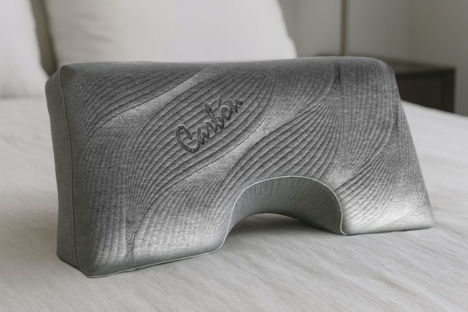 Almohada cervical gris de espuma viscoelástica texturizada sobre una cama, con un recorte arqueado y un logotipo en relieve.