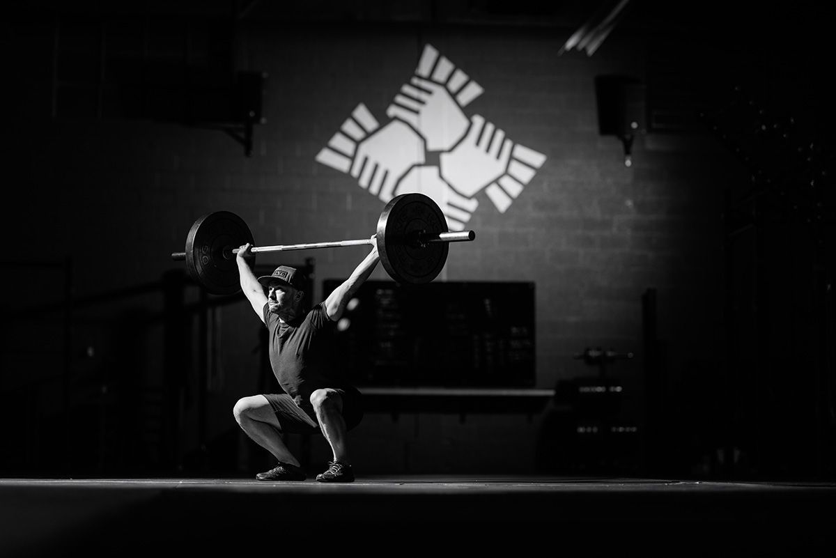 Silhouetted weightlifter squatting under a barbell in a dim gym, with a bright wall emblem behind.