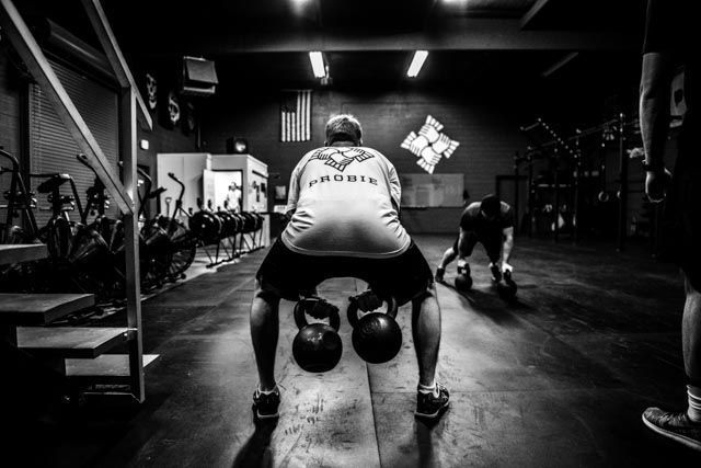 Black-and-white gym scene at The Sect gym with two people doing kettlebell workouts under overhead lights
