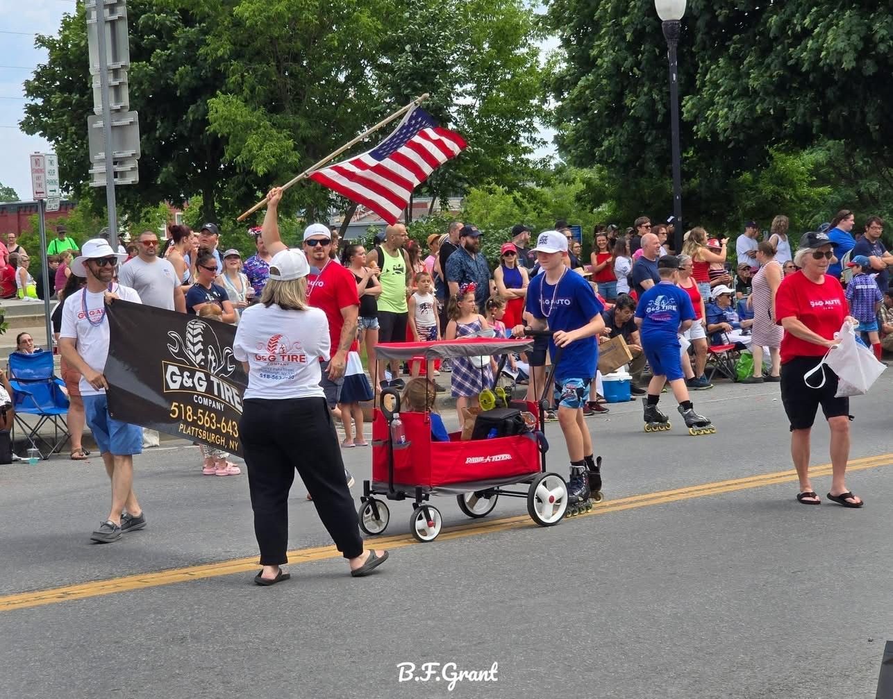 Street parade with marchers and spectators, including an American flag and a red wagon on a tree-lined road