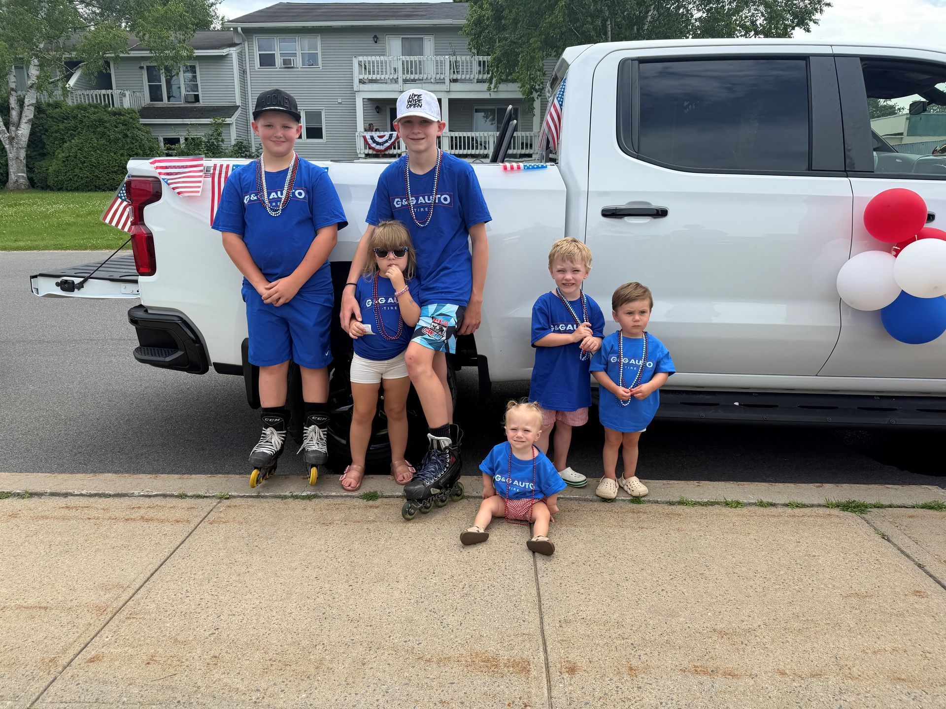 Four children in blue shirts pose beside a white pickup truck decorated with red, white, and blue balloons.