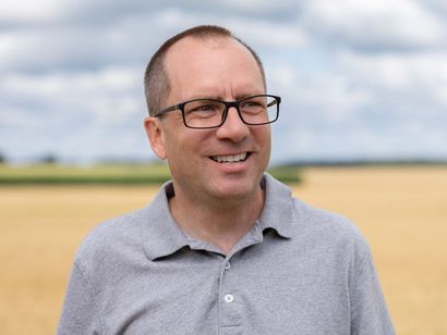 Brad Rocke wearing glasses and a grey shirt is standing in a field.
