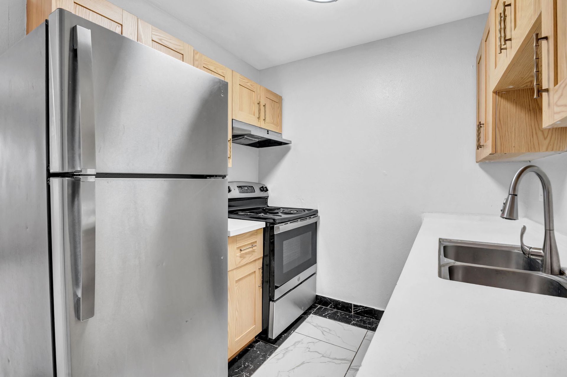 Image of a kitchen with modern tan wood cabinets, and stainless steel appliances with silver hardware and quartz countertops. 