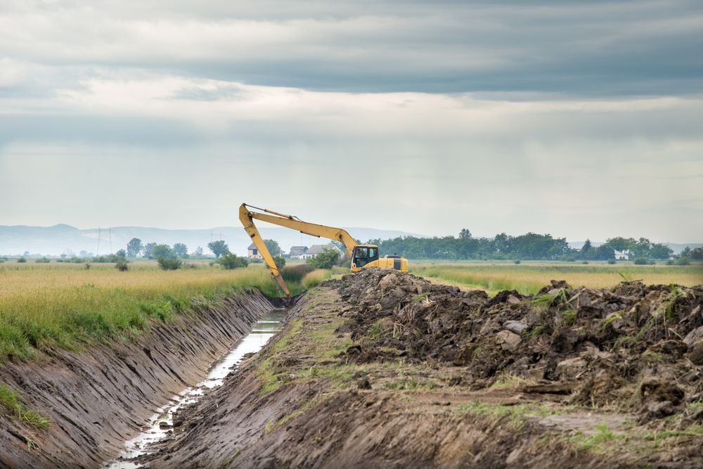 Excavator digging a drainage ditch in a field under a cloudy sky.