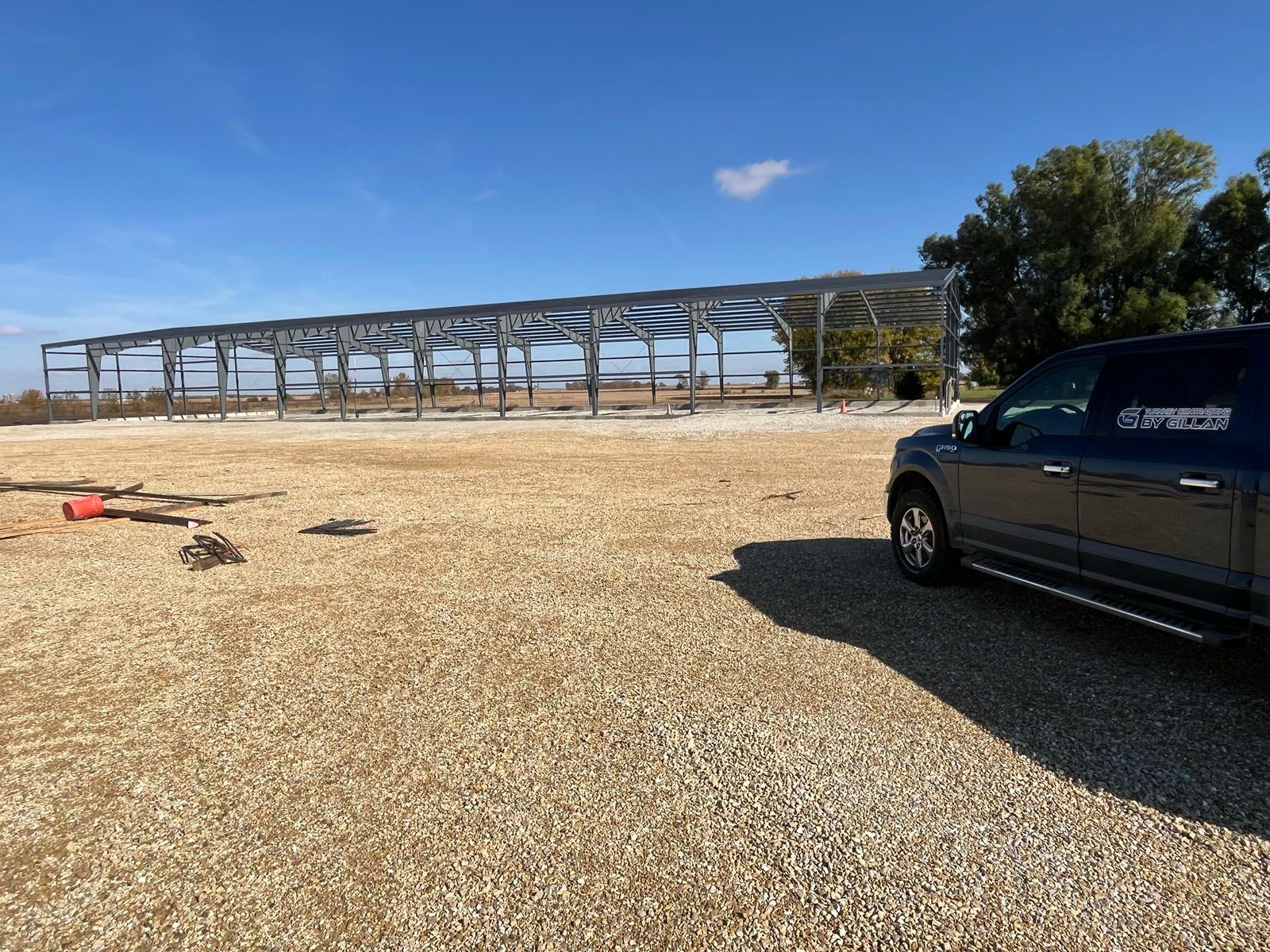 Construction of a steel structure with a truck parked in front on a gravel lot under a blue sky.