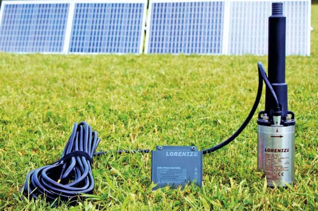 Solar panel equipment on grass: coiled cable, control box, and cylindrical pump beside a solar panel array.