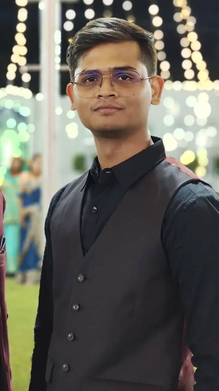 Young man in black shirt and vest wearing glasses, outdoor setting, gazing at camera.