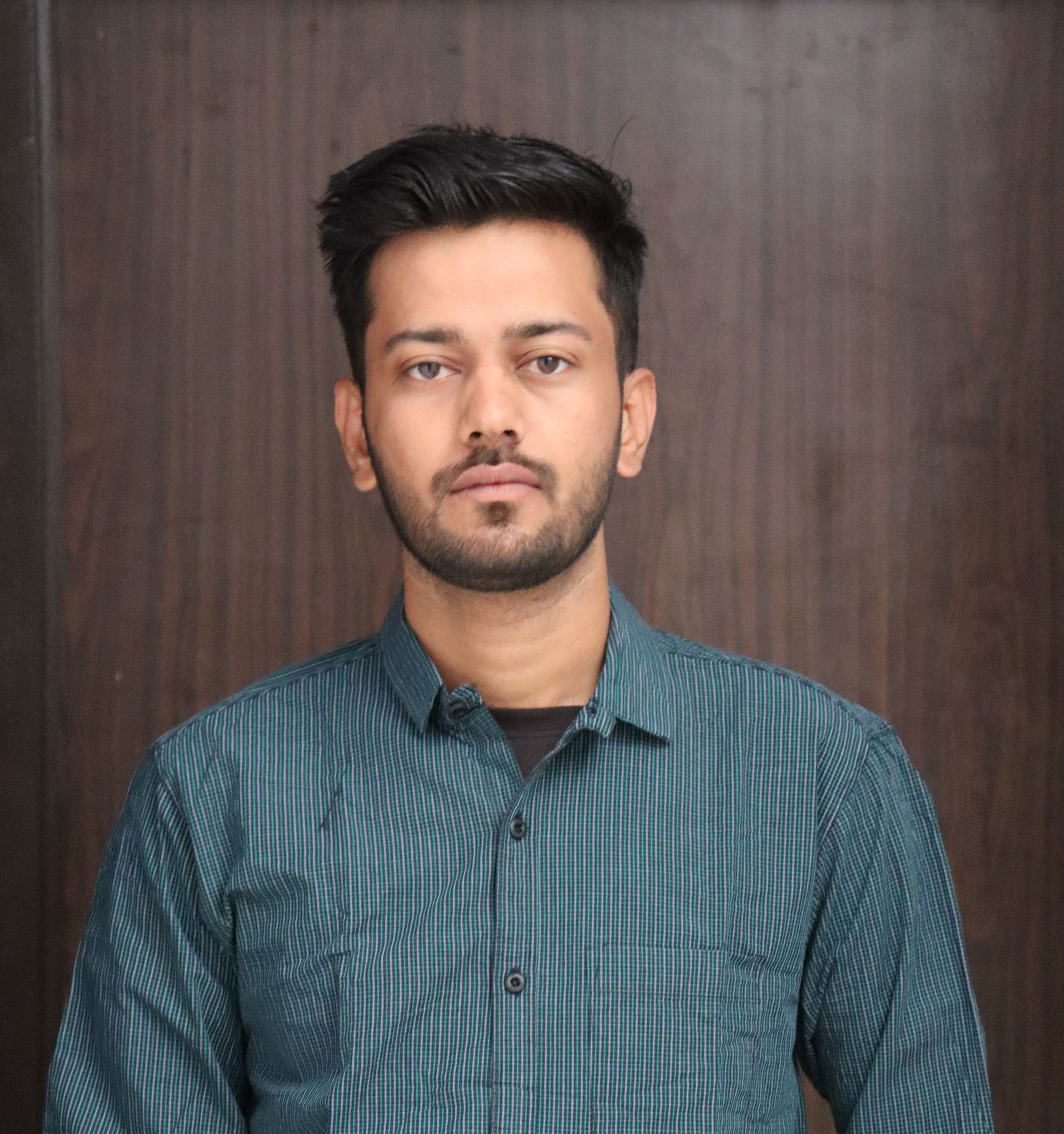 Man with short dark hair and beard, wearing a teal patterned shirt against a brown wood-paneled wall.