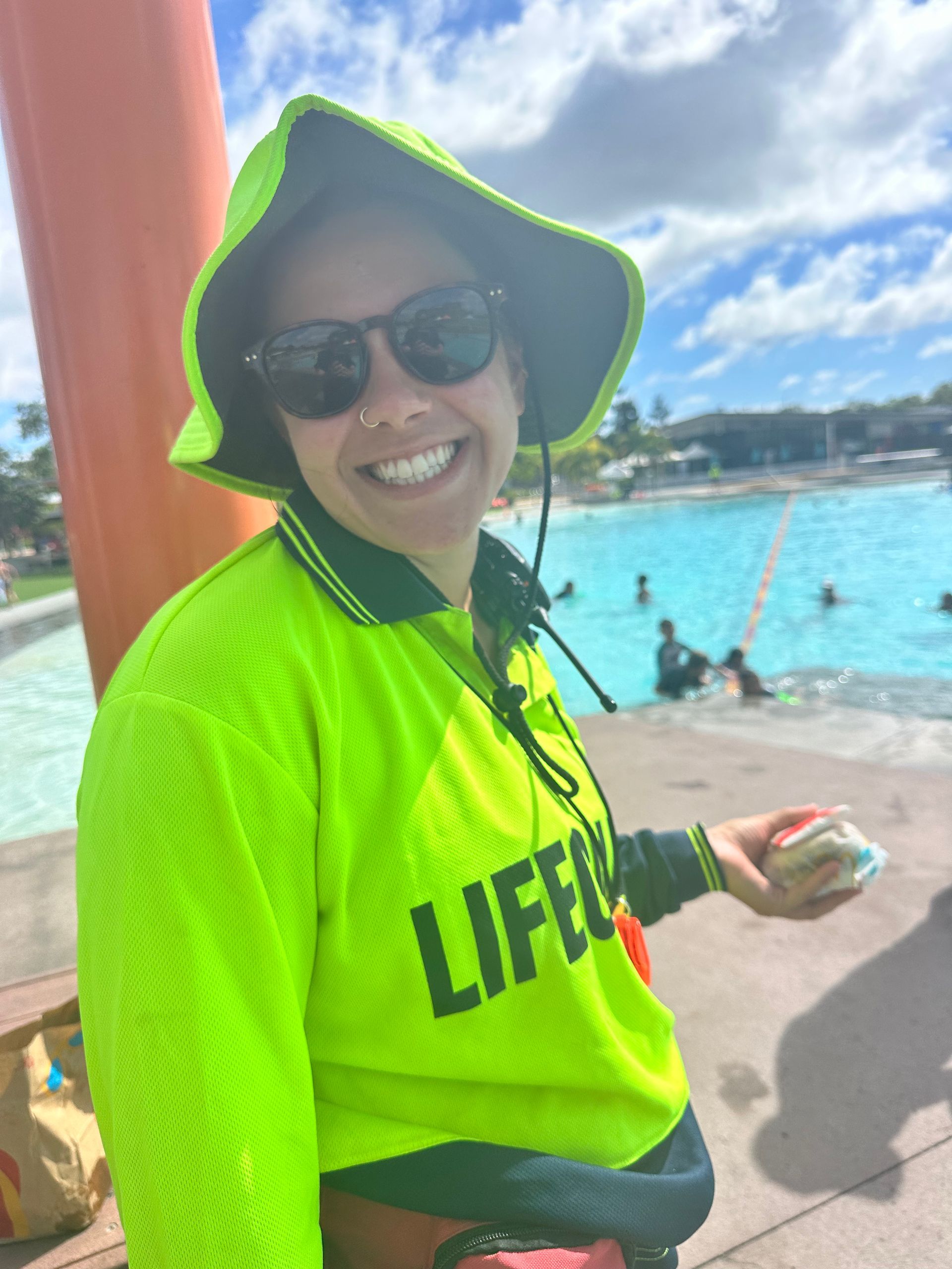 A woman wearing a lifeguard shirt and hat is standing in front of a pool.