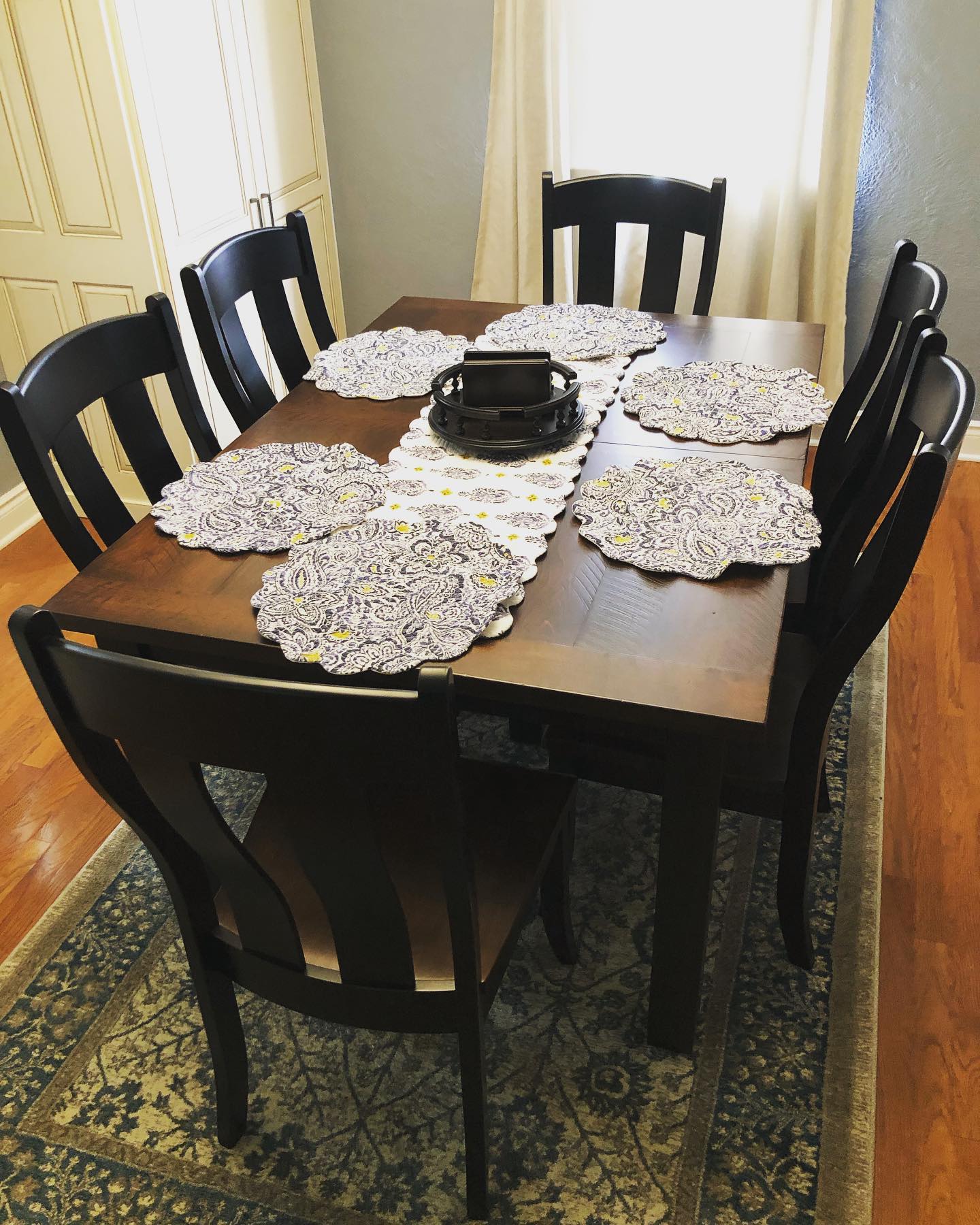 Dining table with six chairs, lace placemats, and a black centerpiece in a bright room