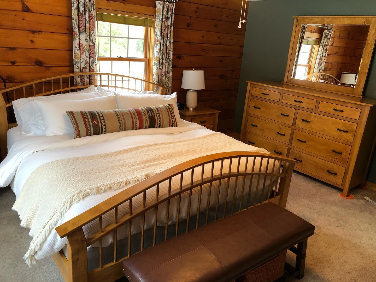 Wood-paneled bedroom with a bed, bench, dresser, lamp, and mirror by a window.