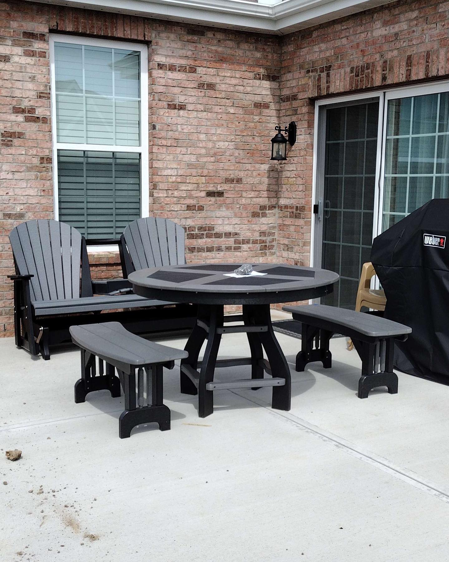 Brick patio with a round black table, benches, Adirondack chairs, and a covered grill beside a house wall.