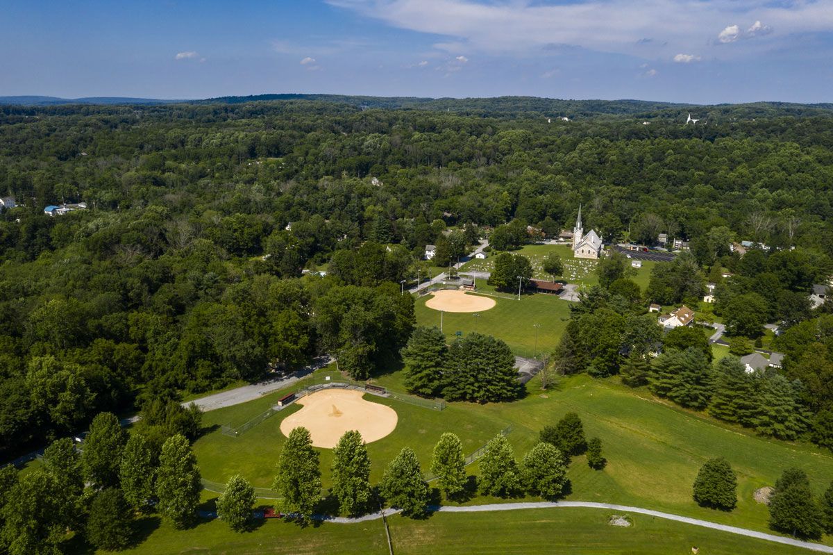 Aerial view of a green park with a baseball diamond, trees, and houses under a blue sky