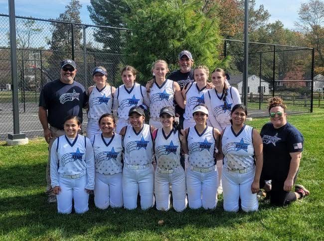 Softball team posing outdoors in white uniforms, grouped on grass near a chain-link fence.