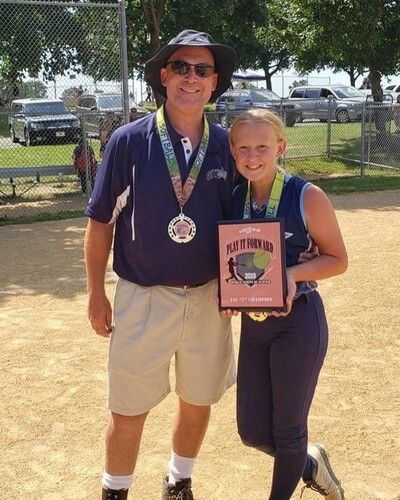 Two people posing outdoors; one holds a pink plaque and both wear medals.