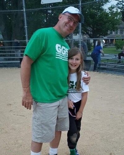 Man in green shirt and girl in white shirt posing together on a baseball field, smiling.