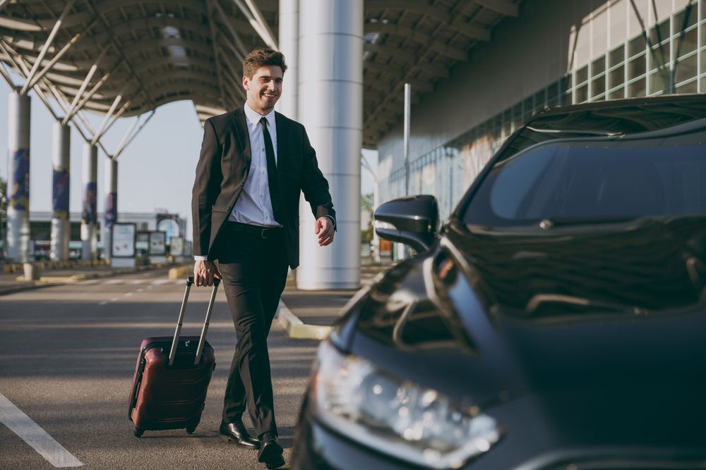 A smiling person in a suit walks toward a black car outside an airport terminal, pulling a small piece of luggage.