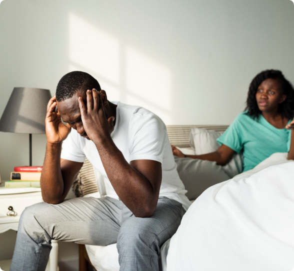 Man with head in hands, sitting on a bed. Woman in background gestures, possibly comforting. Bedroom setting.