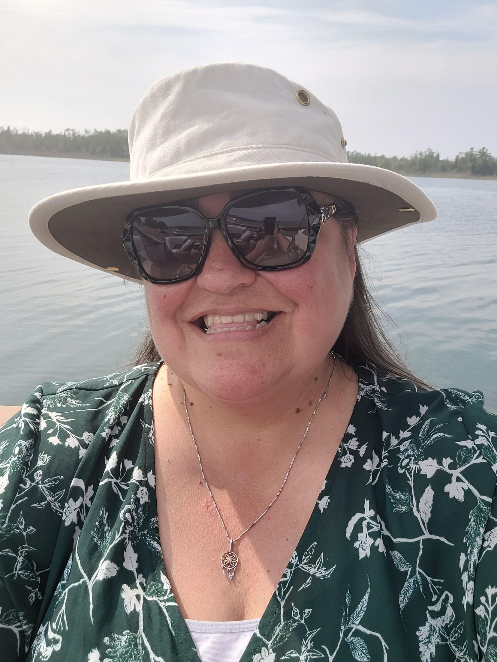 Smiling person in a white sunhat and sunglasses on a boat, with water and shoreline behind them