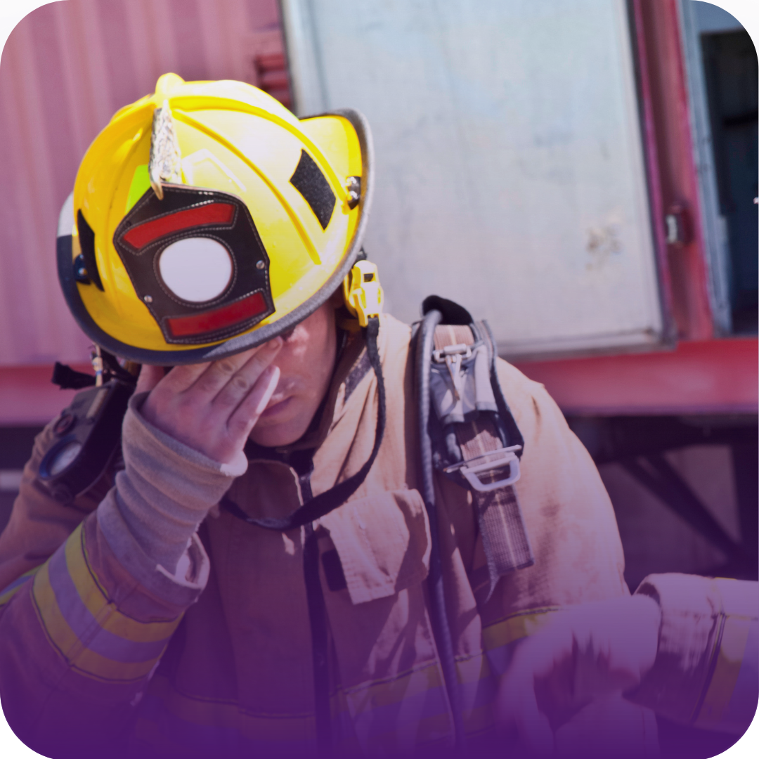 Firefighter wiping face, wearing yellow helmet and gear, near a building.