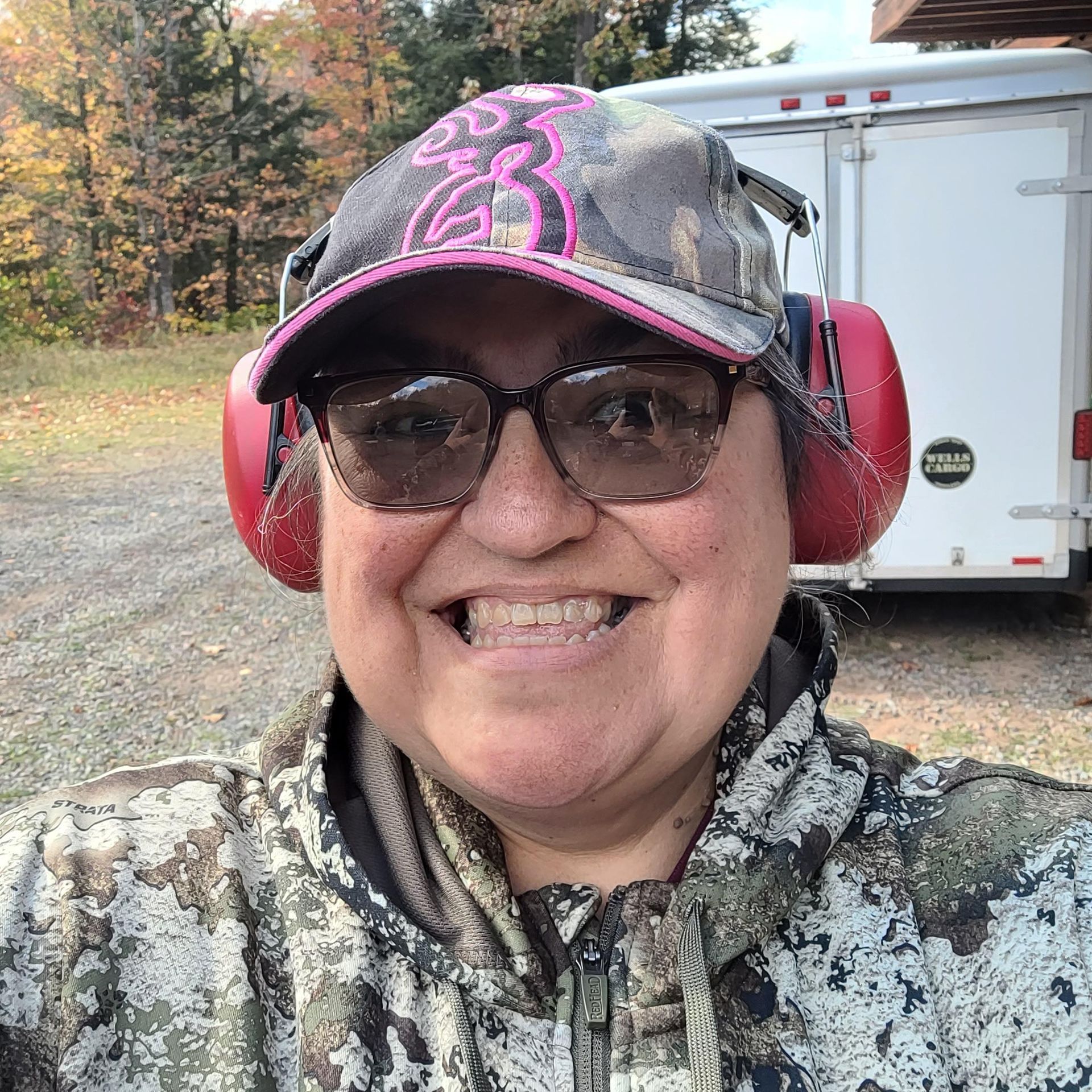 Smiling person in camo jacket and cap with pink headphones, standing outdoors near a trailer.