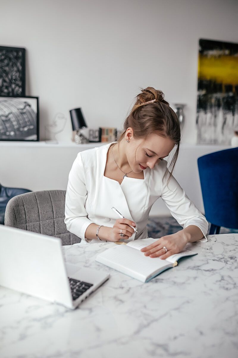Woman in white jacket writing in notebook at a table with laptop, marble surface, and interior setting.