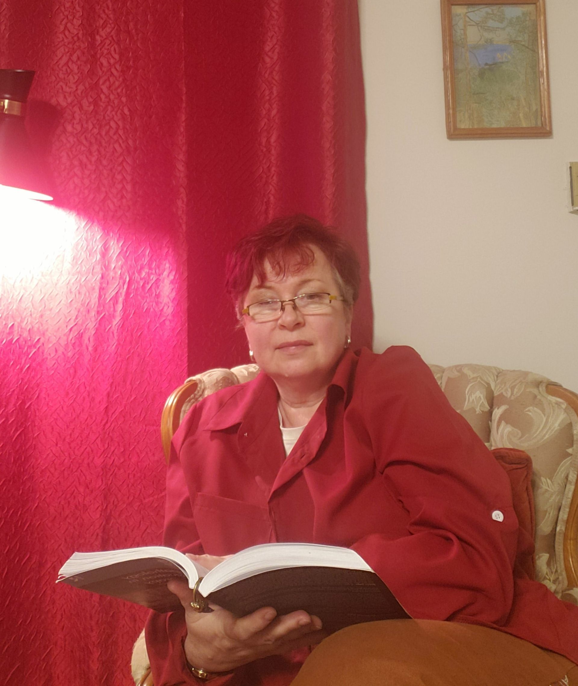 Woman in red jacket seated, reading a book. Red curtains and small framed art in background.
