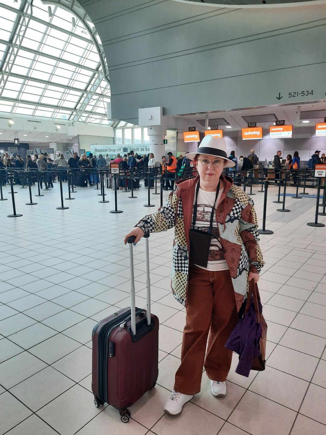Person in a colorful patterned coat and hat at an airport, holding a suitcase and bag, standing near check-in counters.
