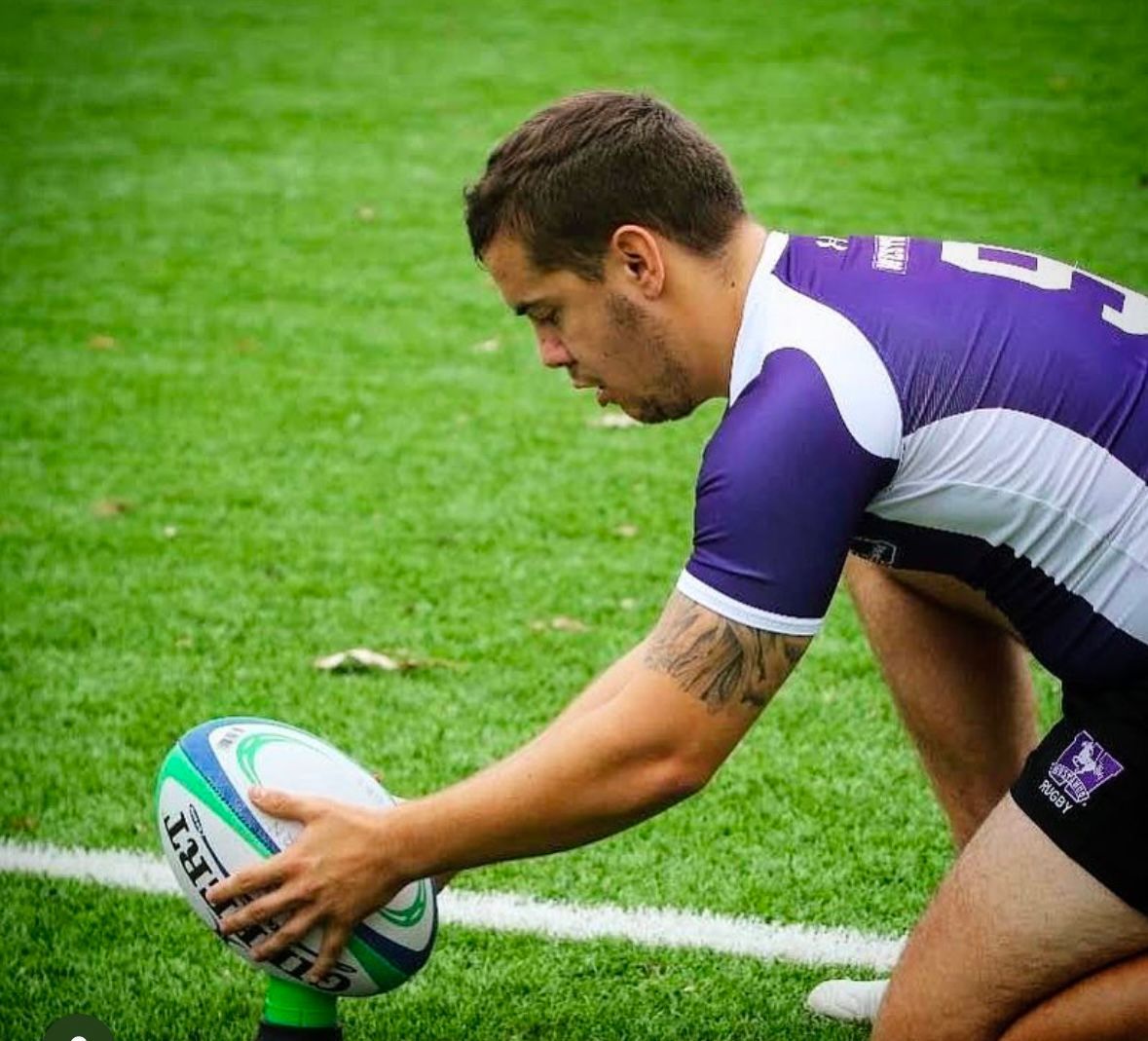Rugby player kneeling, setting up to kick a rugby ball on a green field. Purple and white jersey.
